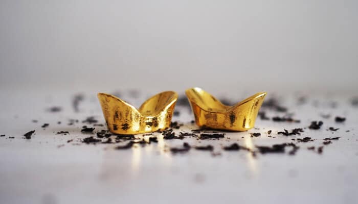 Buying gold bullion Two small gold ingots placed on a white surface with scattered black particles, symbolizing wealth and prosperity associated with buying gold bullion.
