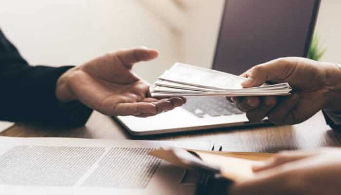 Personal injury compensation A person handing over a stack of cash to another individual across a desk with documents and a laptop, symbolizing the settlement process for personal injury compensation.