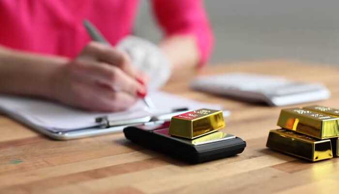 Precious metals Person writing notes at a desk with gold bars and a calculator nearby, symbolizing investment planning and valuation of precious metals.