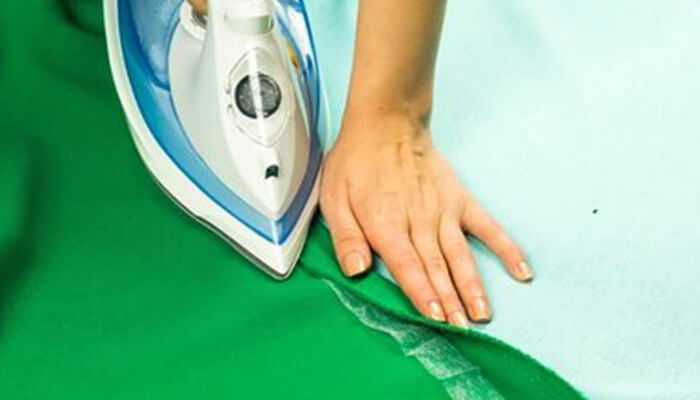 Hemming fabrics without stitching Person using an iron to seal the edge of green fabric, demonstrating a technique for hemming fabrics without stitching.