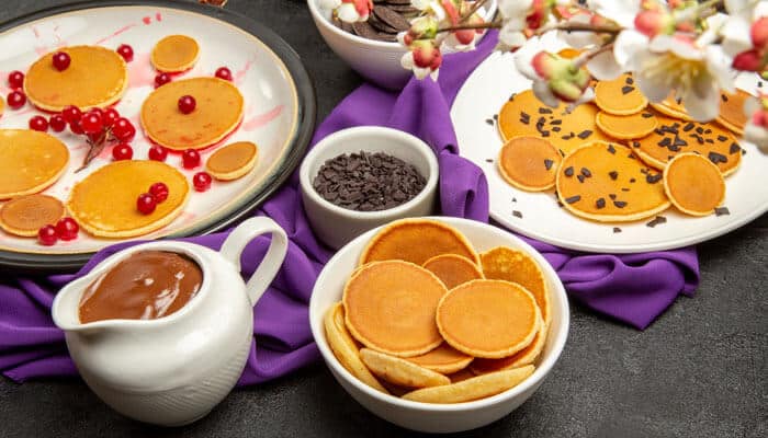 Colorful North Pole breakfast setup featuring mini pancakes, chocolate sauce, red currants, and chocolate chips arranged on plates with a purple cloth and festive floral decor.