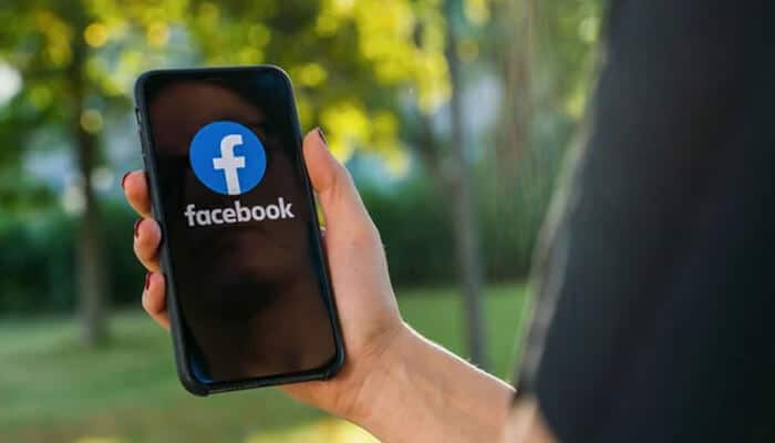 Adjust profile and photo visibility - tycoonstory | tycoonstory media Person holding a smartphone displaying the facebook logo app screen outdoors, representing checking or updating facebook profile visibility.