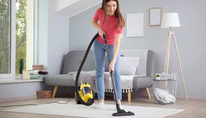 Sustainability as a design imperative - tycoonstory | tycoonstory media A young woman using a yellow modern vacuum to clean a bright living room carpet, highlighting efficiency and everyday innovation.