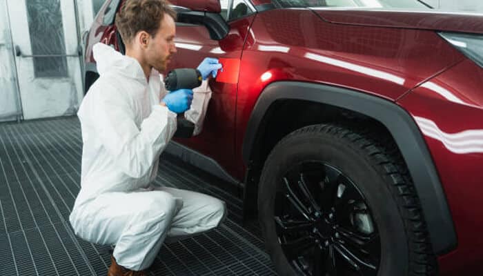 Seasonal car care Auto technician in protective coveralls inspecting and polishing the side panel of a red suv under workshop lighting, representing seasonal car care focused on maintaining paint quality and vehicle condition year-round.