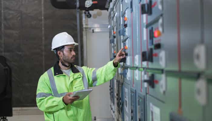 Key design features of expertly engineered switchgear | tycoonstory media A technician wearing a hard hat and high-visibility jacket inspecting metal enclosed switchgear panels inside an industrial facility.