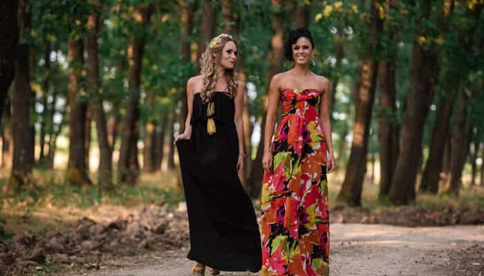 Designer maxi dress Two women walking along a forest path, each wearing a colorful designer maxi dress—one in a black dress with tassels and the other in a vibrant multicolored pattern.
