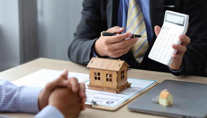 Home loan interest rate A financial advisor discusses numbers on a calculator with a client, with a small wooden house model on the desk, illustrating how a home loan interest rate is calculated.