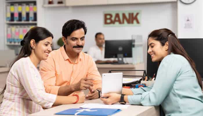 Credit guarantee scheme A bank representative shows information on a tablet to a smiling couple seated at a desk, illustrating customer support and financial assistance under the credit guarantee scheme.