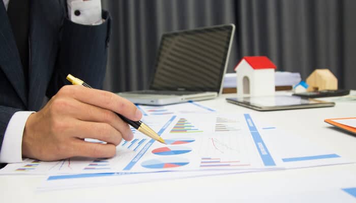 Comparing returns funds versus real estate | tycoonstory media A person reviewing charts and graphs related to investments, possibly discussing the financial returns of funds versus real estate for u. S. Golden visa applicants. A laptop and model house are visible in the background, indicating a professional setting.