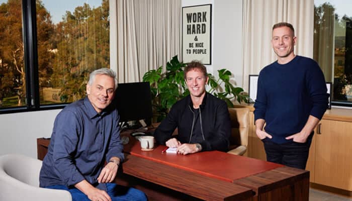 Cowherds | tycoonstory media Colin cowherd (left) with two colleagues in an office, posing around a desk. A sign in the background reads "work hard & be nice to people.