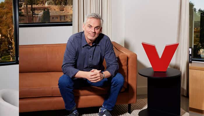 Volume coincides | tycoonstory media Colin cowherd’s relaxed pose on a couch next to a red'V' symbol representing The Volume, set in a modern office environment.