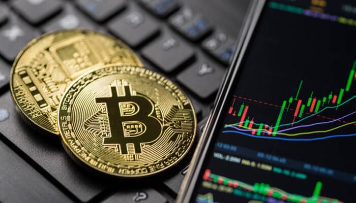 2 | tycoonstory media Bitcoin coins on a keyboard next to a smartphone showing market trends. A reminder to invest in bitcoin today