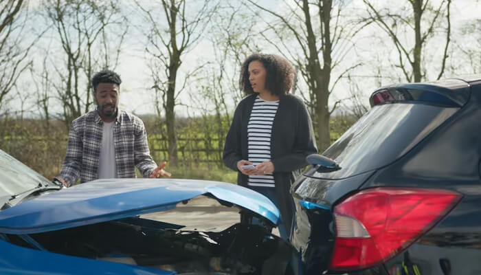Avoid admitting fault | tycoonstory media A man and a woman standing by their cars after a car collision, discussing the accident. The blue car has visible damage to the rear, while the other car is intact.
