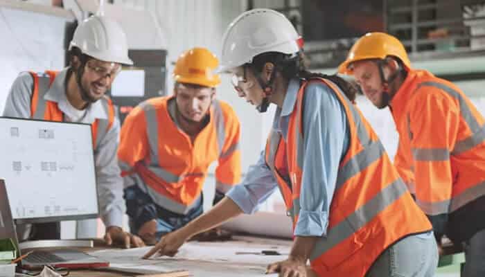 How to handle the massive amount of detail in a project 1 | tycoonstory media A group of construction workers in safety gear discusses plans, emphasizing the importance of using construction specification software to manage project details.