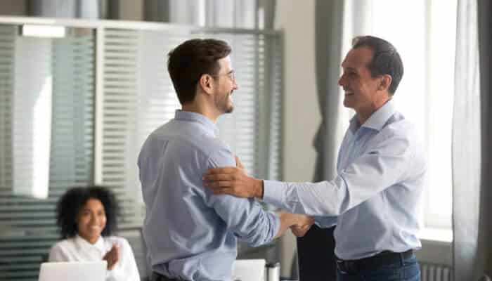 Recognition and appreciation systems | tycoonstory media Two professionals in a modern office environment shaking hands, demonstrating collaboration in the modern workforce, with a colleague smiling in the background.