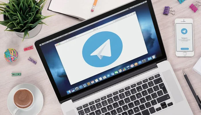 Telegram web version vs mobile app | tycoonstory media Telegram displayed on a laptop and mobile phone, showing the app’s interface in a workspace with a cup of coffee, plant, and stationery items.