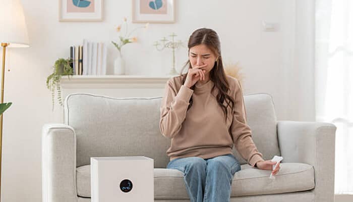 The impact of unhealthy air on lung health | tycoonstory media Woman sitting on a couch, holding a tissue and covering her nose, experiencing discomfort due to poor air quality. This highlights the health risks of sewer exposure and the impact of unhealthy air on lung health.