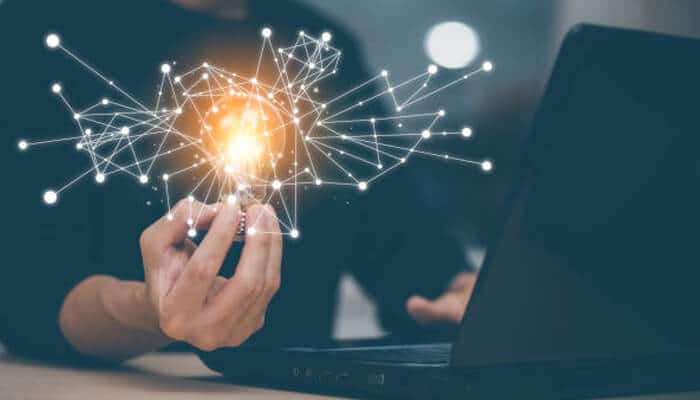 Use your network to spark ideas | tycoonstory media Person holding a glowing digital lightbulb with network connections near a laptop, symbolizing innovative ideas and strategy to start a business in the digital age.