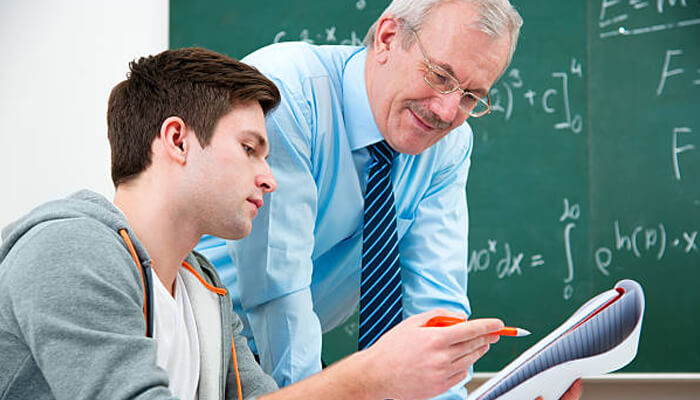 Bring your work and notes to each session | tycoonstory media A math tutor helps a student with their work, pointing to a notebook filled with notes while standing in front of a chalkboard with mathematical equations.