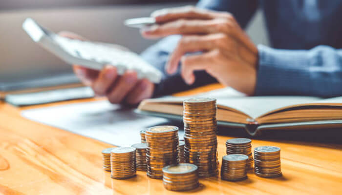 Build a practical plan for money and operations | tycoonstory media A person calculating finances with a calculator, surrounded by stacks of coins and an open book, reflecting the financial planning involved in launching your first business.