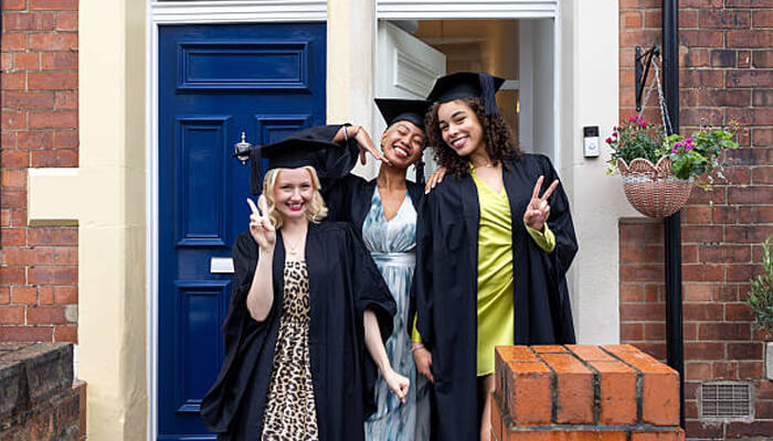 Choose a shape that works under a gown 1 | tycoonstory media Three graduates celebrating with peace signs in front of a house. A perfect dress for graduation events, like these stylish outfits, ensures comfort and style.