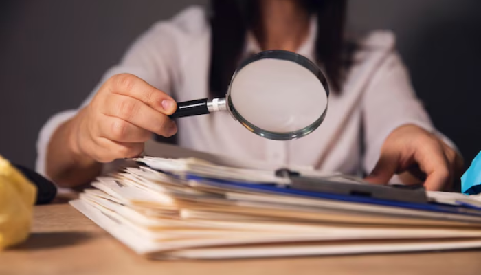 How attorneys evaluate claims of hostile work environments | tycoonstory media A person using a magnifying glass to review documents, symbolizing how attorneys evaluate claims of hostile work environments by carefully examining evidence and details.