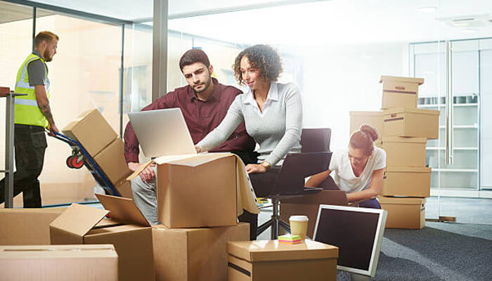 Protect systems records and physical assets | tycoonstory media A team of professionals working on tasks while surrounded by moving boxes, illustrating the process of relocating your startup office to a new, more efficient space.