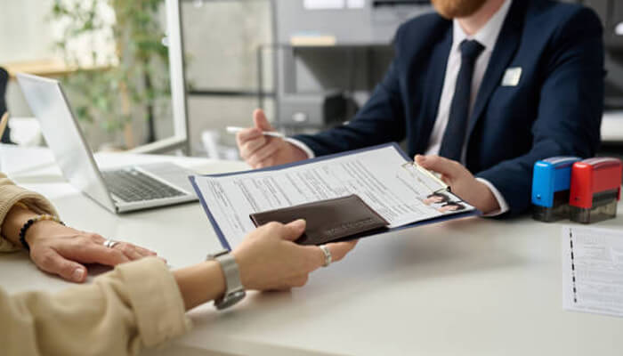 Submitting the application | tycoonstory media A person submitting documents to an agent, possibly for a partner visa subclass 820 application, with a laptop and stamping tools visible on the desk.
