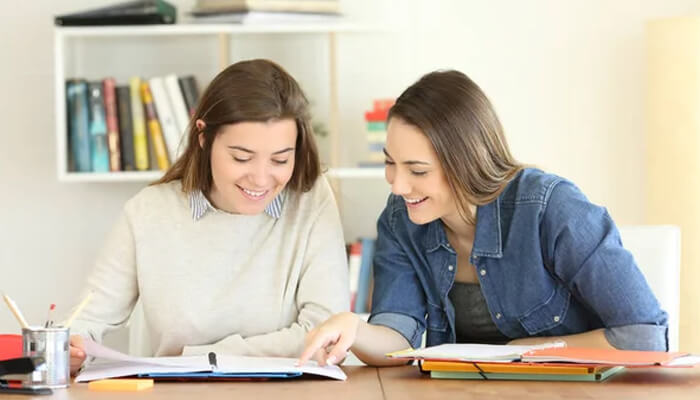 Teaching others as a study strategy | tycoonstory media Two women smiling and pointing at study materials on a table, using collaboration as part of the best study methods to learn together.