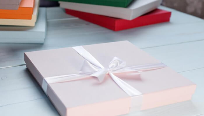Custom book boxes | tycoonstory media Pink gift box tied with a white ribbon on a light blue table, with colorful books blurred in the background.