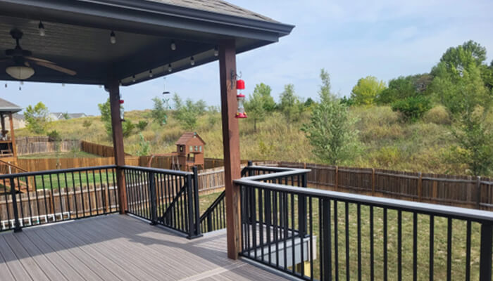 E 3 | tycoonstory media Covered outdoor deck with railings and hanging bird feeder overlooking a grassy backyard and trees in the distance, with a ceiling fan overhead.