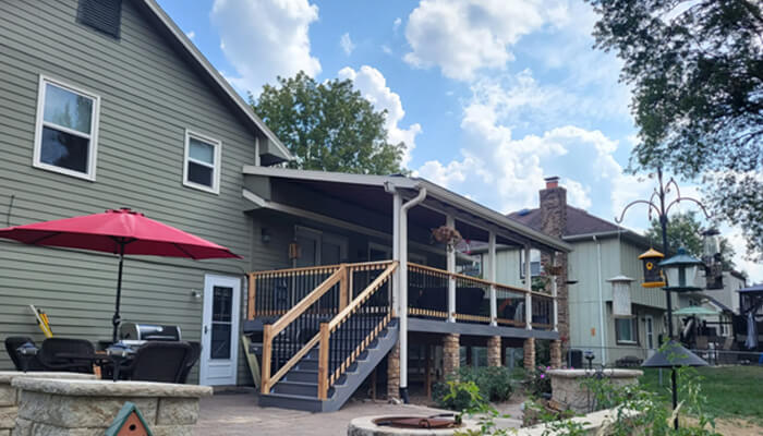 R 2 | tycoonstory media Backyard scene with a gray-green house, a wooden deck and stairs, black railing, a red patio umbrella, and outdoor seating.