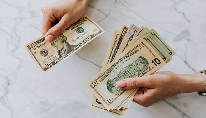 Asset-backed and secured lending | tycoonstory media Two hands exchange us dollar bills on a light marble surface, with one hand counting and the other ready to hand over a stack of cash.