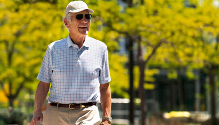 E 4 | tycoonstory media Older man wearing a white cap, sunglasses, and a light plaid shirt walks outdoors in a sunny park.