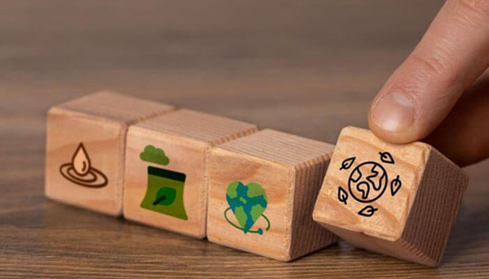 How smart electrical systems support esg and regulatory goals | tycoonstory media Wooden blocks with eco-icons—candle, green bag, globe with a heart—lined up on a table, with the last block being touched by a finger.