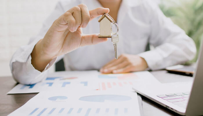 It controls | tycoonstory media Person holds a small house key on a keyring above documents and a laptop, suggesting real estate or mortgage planning.