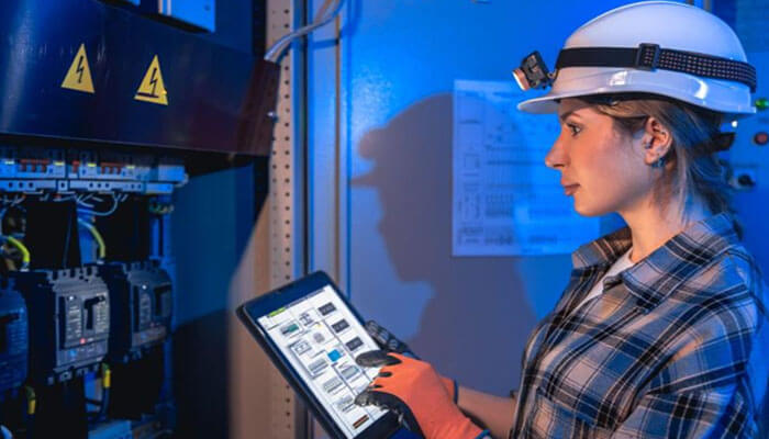 Key technologies behind smart electrical systems | tycoonstory media Female technician in a plaid shirt and hard hat uses a tablet to inspect an electrical panel in an industrial cabinet room.