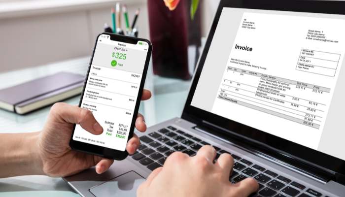 Same day invoicing | tycoonstory media Close-up of a hand holding a smartphone showing a 5 invoice, while a laptop screen displays a printed invoice.