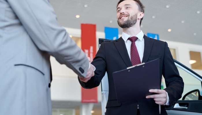 Standard methods for determining business worth | tycoonstory media Two businessmen shake hands in a car showroom; one holds a clipboard and wears a suit, car in the background.