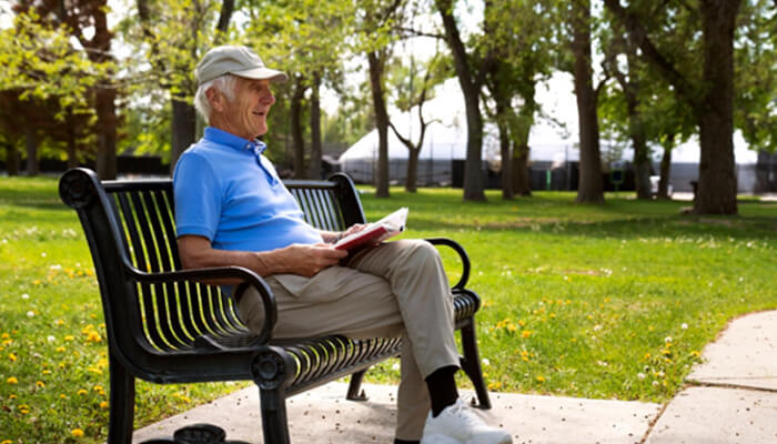 T 2 | tycoonstory media Older man in a blue polo reads a magazine while sitting on a park bench on a sunny day in a green, tree-lined park