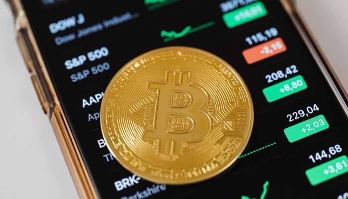 The number on the screen is not the number in your wallet | tycoonstory media Bitcoin coin resting on a smartphone screen displaying crypto prices and green/red indicators on a trading app