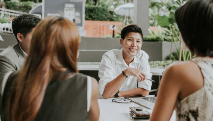 The wellness industry is built on habit | tycoonstory media Smiling man in a white shirt talks with three coworkers at a round table in a modern office setting.