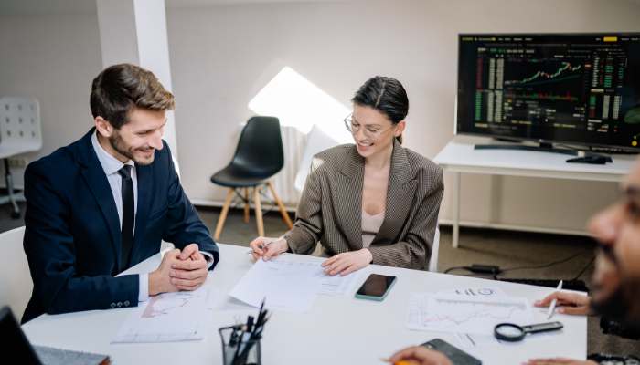 Unified visibility across teams | tycoonstory media Two professionals in a meeting at a white table reviewing documents, with a monitor displaying charts in the background.