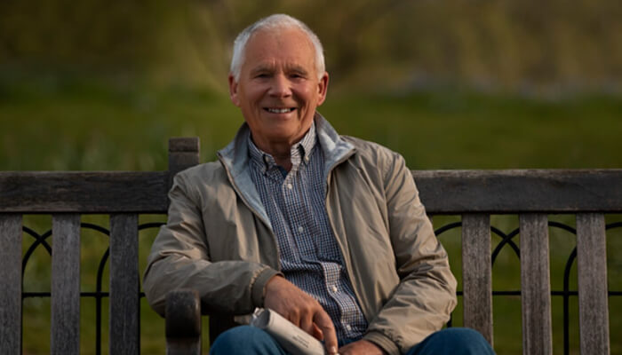 W 3 | tycoonstory media Smiling older man sits on a wooden park bench outdoors, wearing a beige jacket and blue checkered shirt.