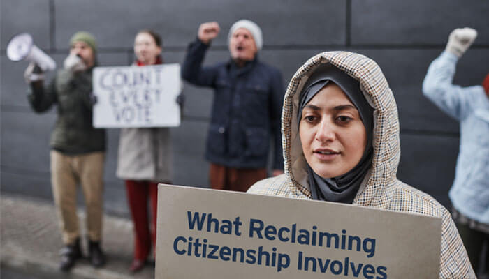 What reclaiming citizenship involves - tycoonstory | tycoonstory media Protester in a hijab and plaid coat holds a sign that reads 'what reclaiming citizenship involves' as other demonstrators protest in the background.