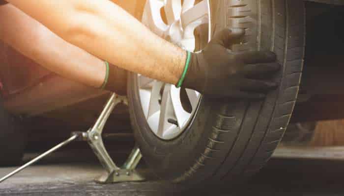 5 wheel and tire upgrade mistakes to avoid 1 | tycoonstory media Person wearing gloves changing a car tire, using a floor jack to lift the vehicle.
