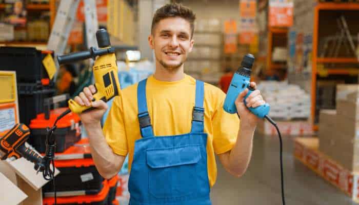 7 essential safety tips for diy enthusiasts operating high-voltage power tools 1 | tycoonstory media Male hardware store worker in yellow shirt and blue overalls holding a yellow drill and a blue power tool, smiling at the camera in a workshop aisle.