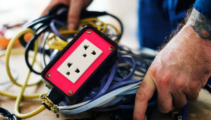7 outdoor extension cord storage ideas for safer power management 3 | tycoonstory media Person’s hands among tangled extension cords and a red electrical outlet box with a standard two-socket outlet on the floor, suggesting wiring work.