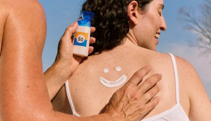7 tips for selecting the best body sunscreen for sensitive skin 1 | tycoonstory media Person applies spf 50 sunscreen on another person’s back at the beach; a sunscreen smiley is drawn on the shoulder.