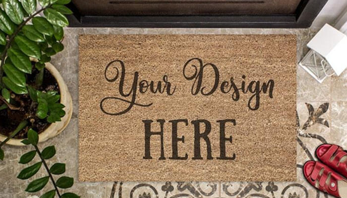 A personalized doormat or yard sign - tycoonstory | tycoonstory media Doormat on a doorstep that says 'your design here' in script and bold letters, with a plant and sandals nearby.